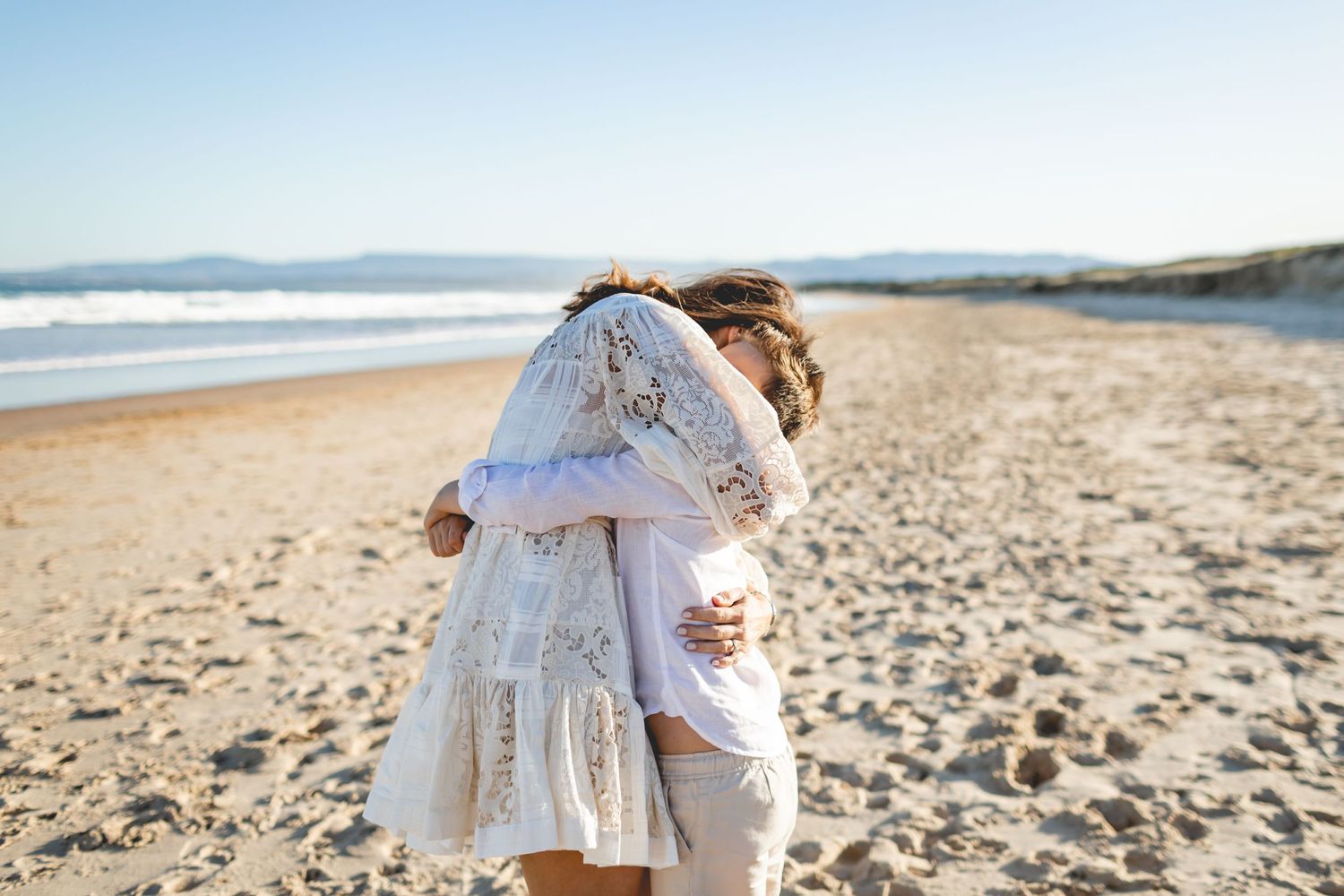 A couple embraces in white outfits on a sunny beach with waves and sand dunes in the background.