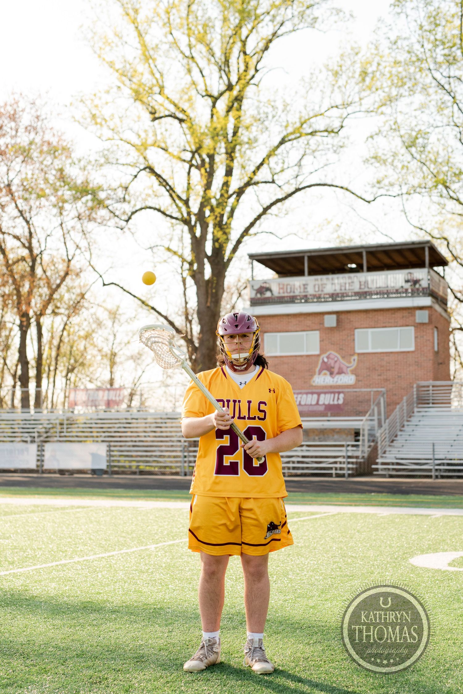Athlete in yellow Bulls jersey number 28 tosses a ball on sports field with bleachers and trees.
