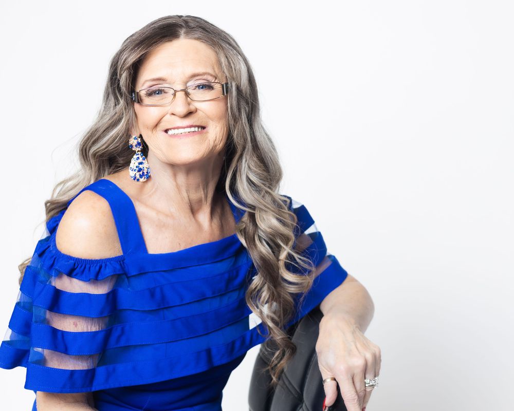 Older lady in blue off the shoulder top posing with her arm over the back of a chair in a white room