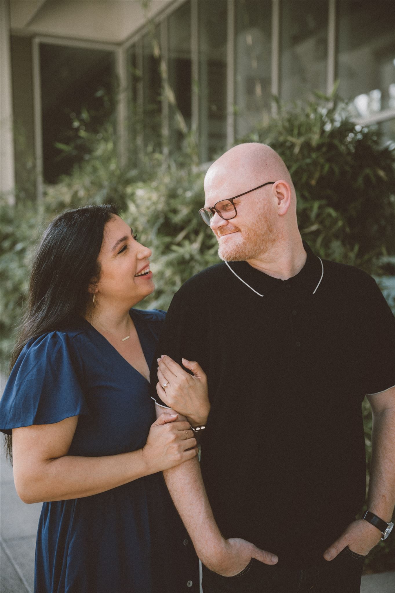 A couple in casual wear share an intimate moment in front of a modern building exterior.
