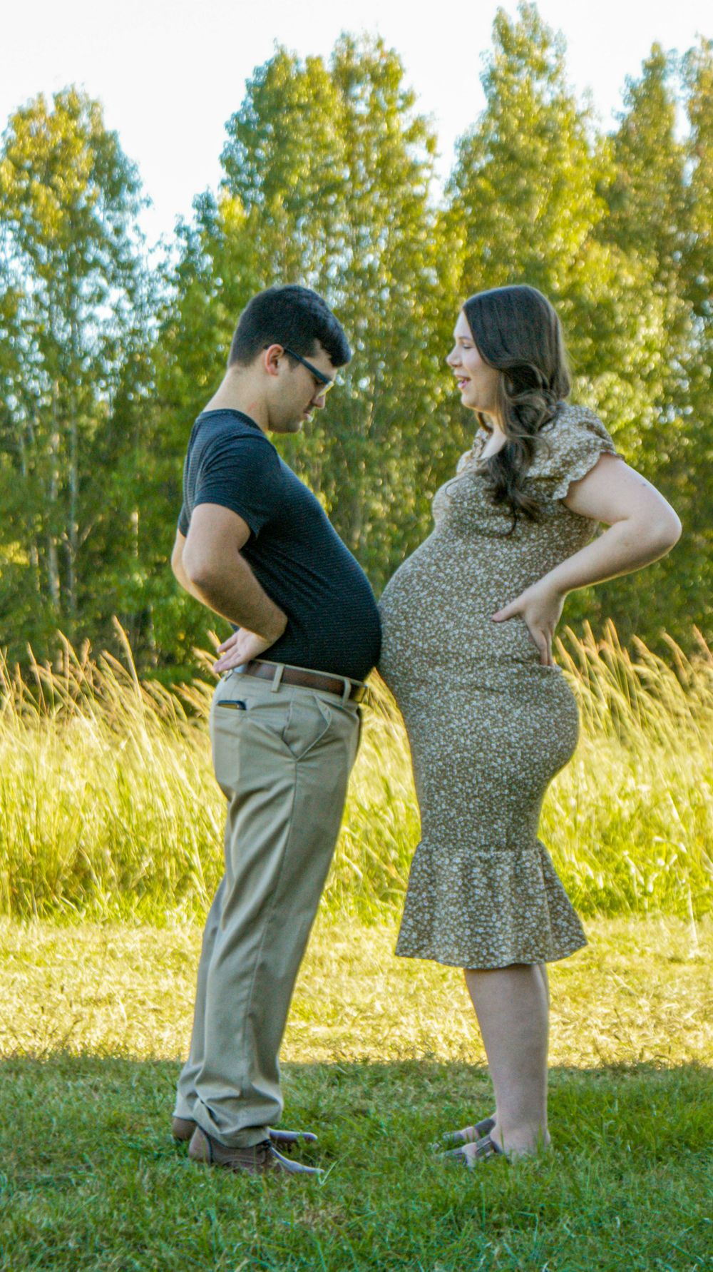 A playful maternity photo with a couple comparing their bellies in a sunny field.