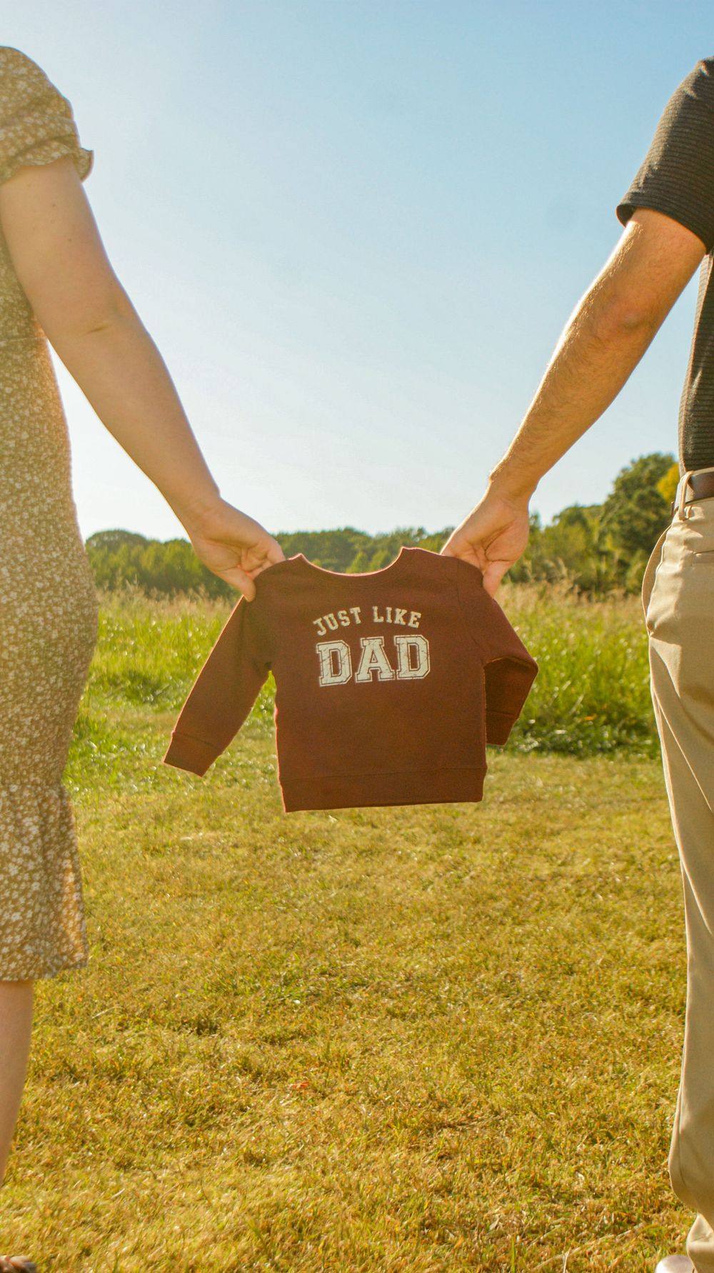 A couple holds up a tiny brown sweatshirt with 'Just Like Dad' text in a sunny meadow.