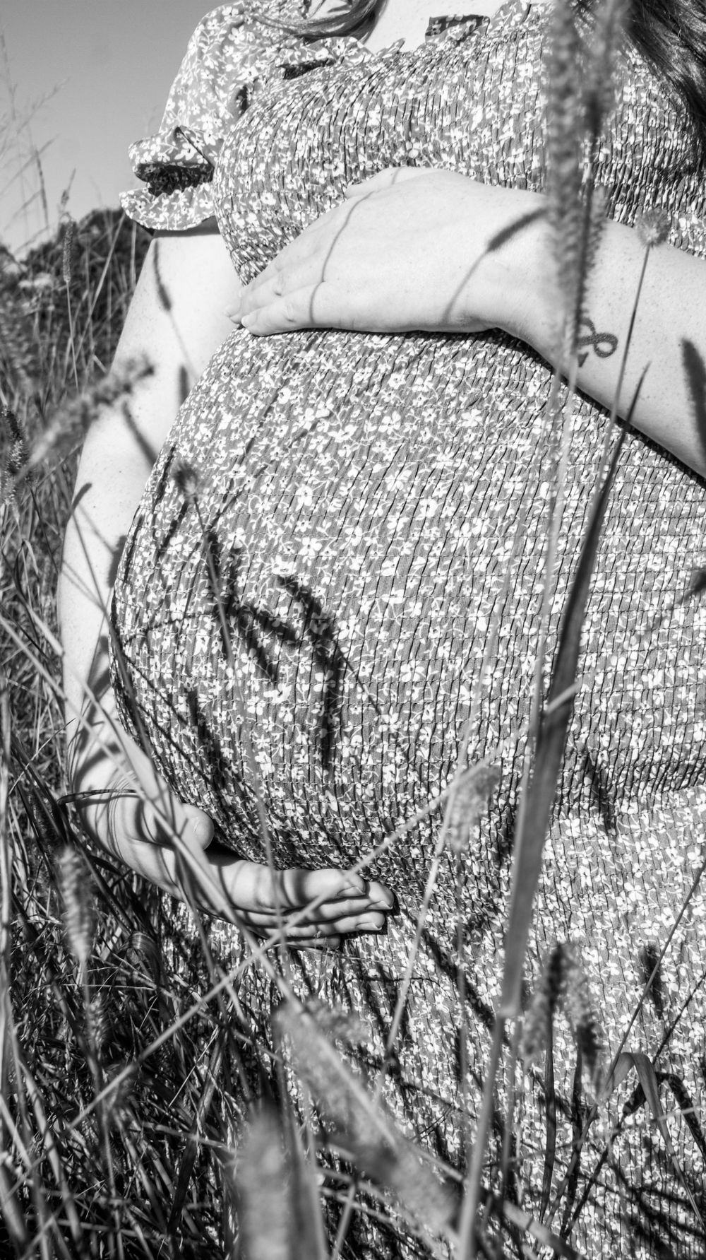 Black and white closeup of a pregnant belly in a floral dress against tall grass.