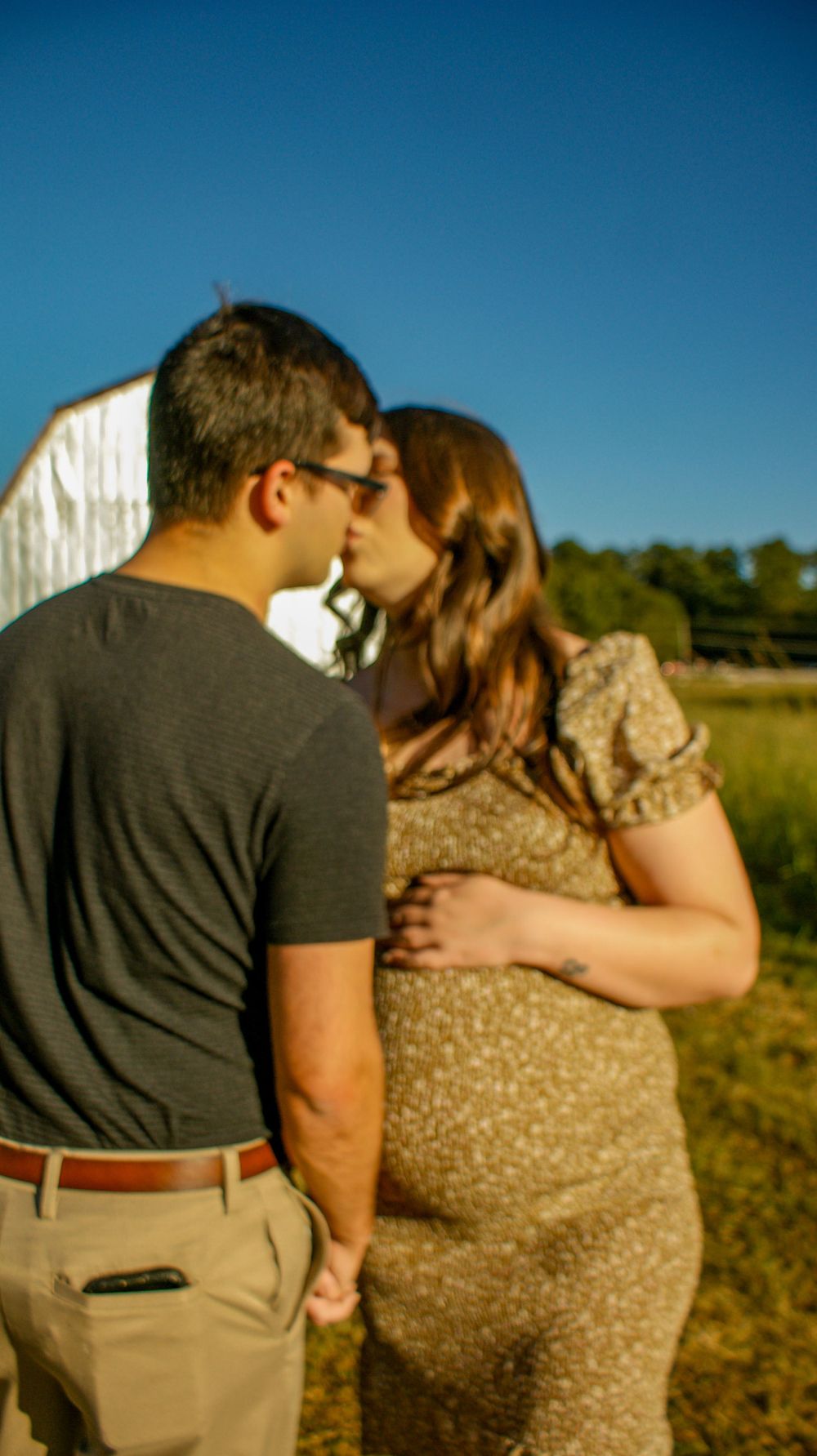 A tender maternity moment captured against a bright blue sky and rural backdrop.