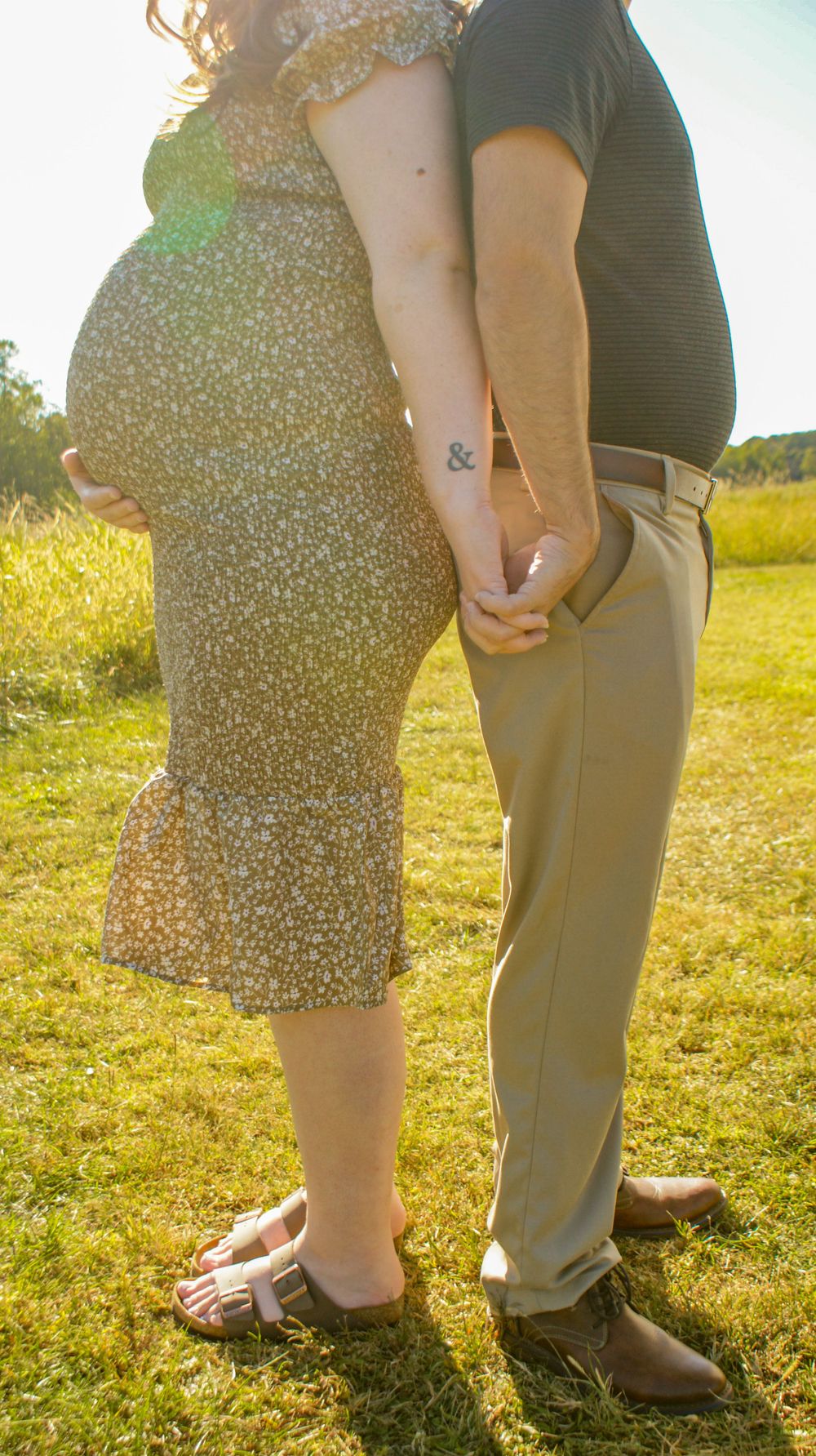 A couple holding hands in a sunlit field, with the woman wearing a floral dress and standing close together.