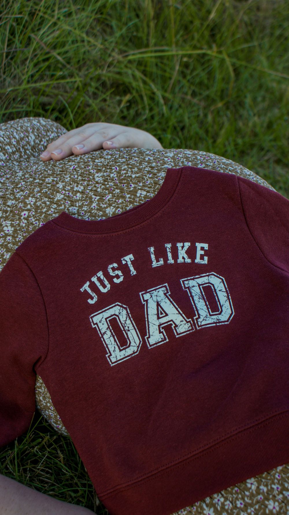 A burgundy baby sweatshirt with 'Just Like Dad' text rests on a floral dress.