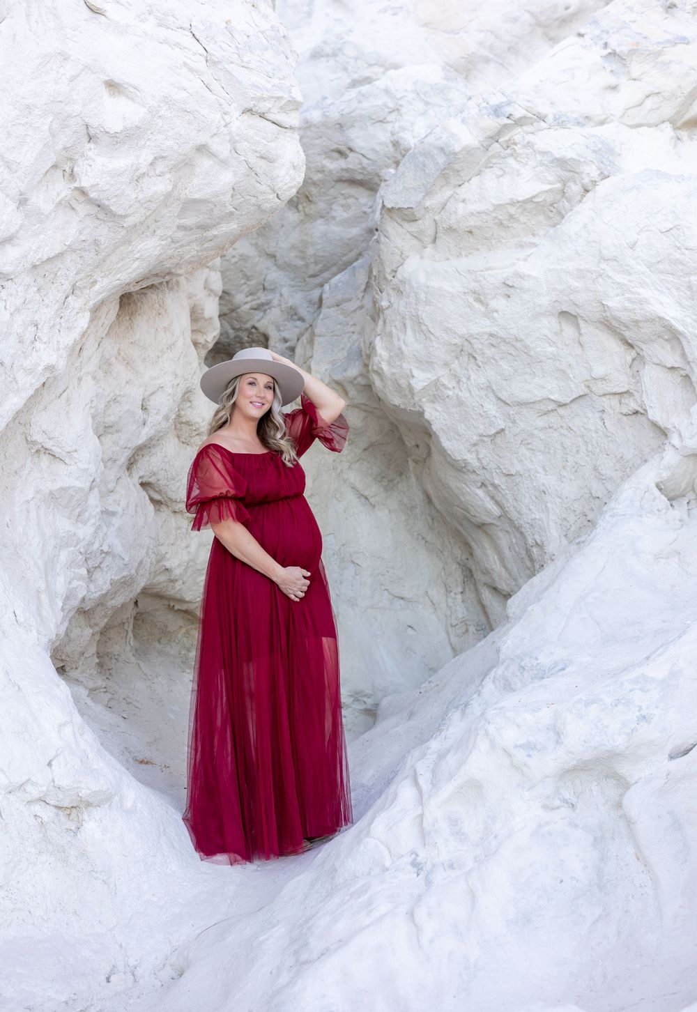 Maternity shoot featuring woman in flowing red dress against white stone backdrop.