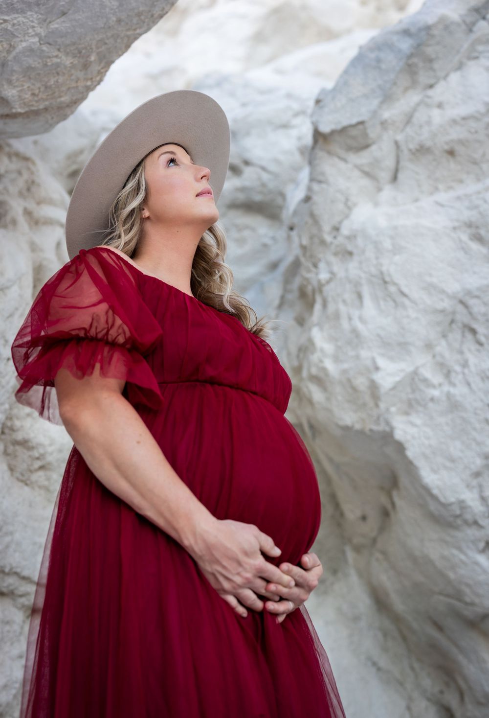 Pregnant woman in burgundy dress and hat poses against white rock formation.