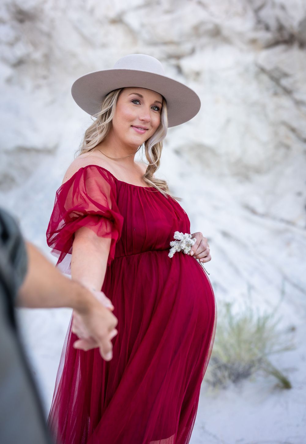 Expectant mother in red dress and hat smiles during outdoor photoshoot.