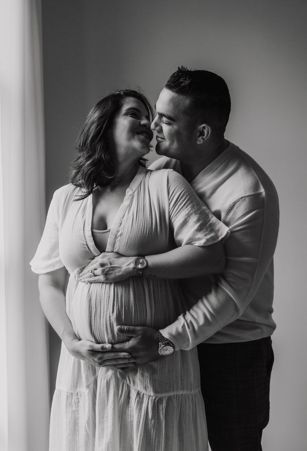 Intimate black and white maternity photo of couple embracing by window.