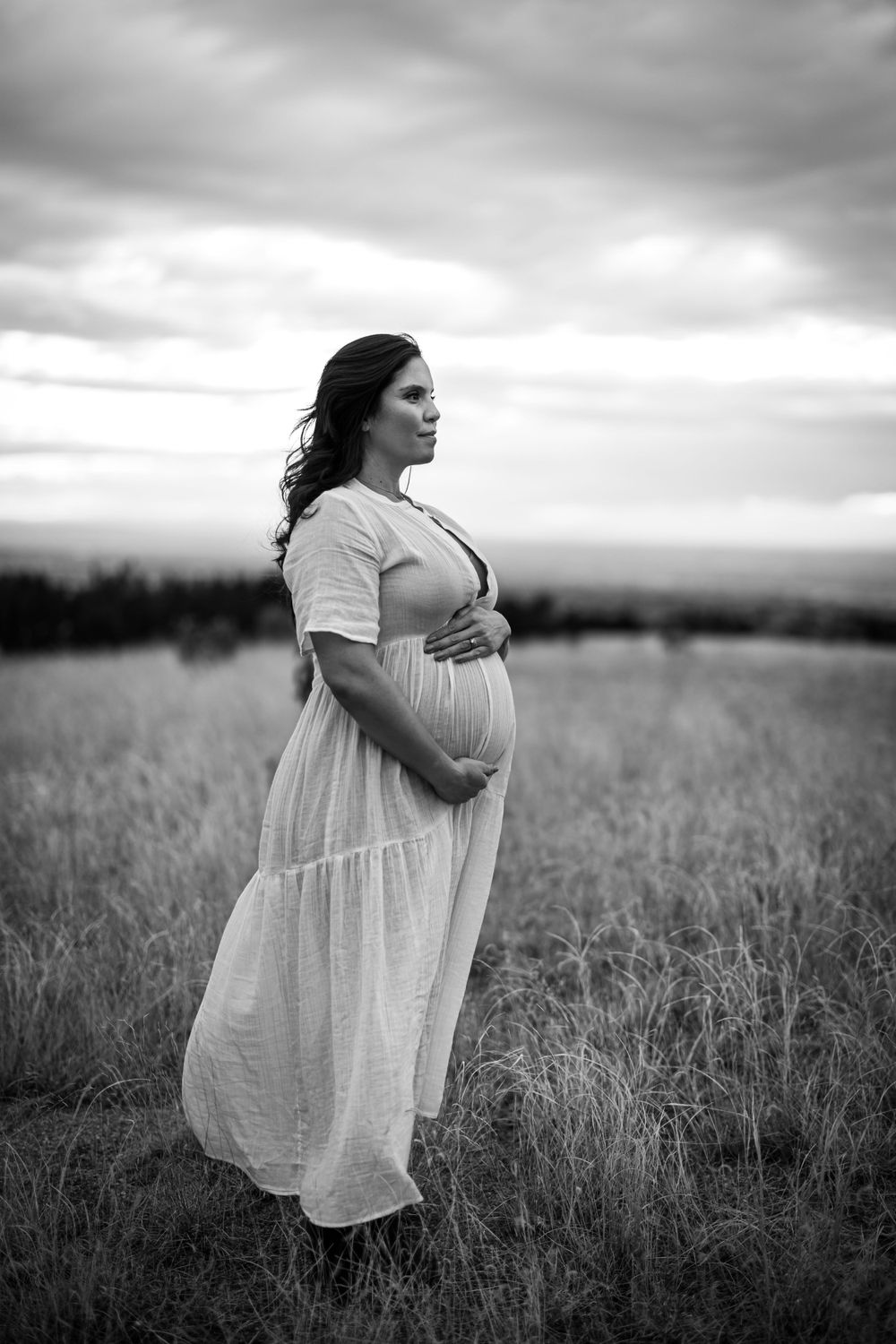 Black and white landscape portrait of pregnant woman in flowing dress standing in field.