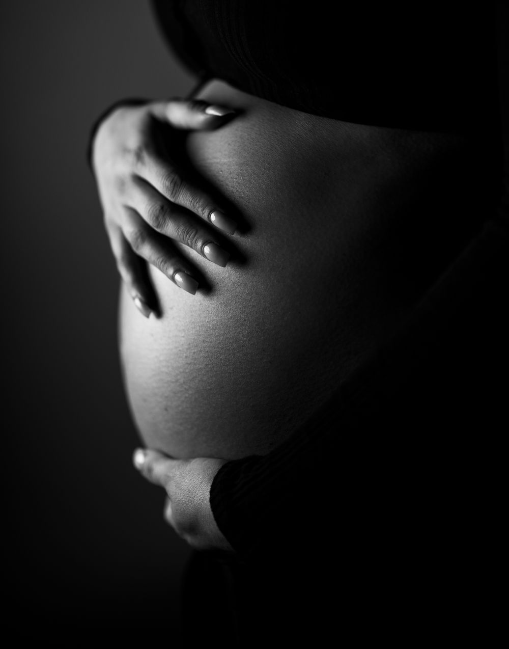 Artistic black and white close-up of hands cradling a pregnant belly in shadow.