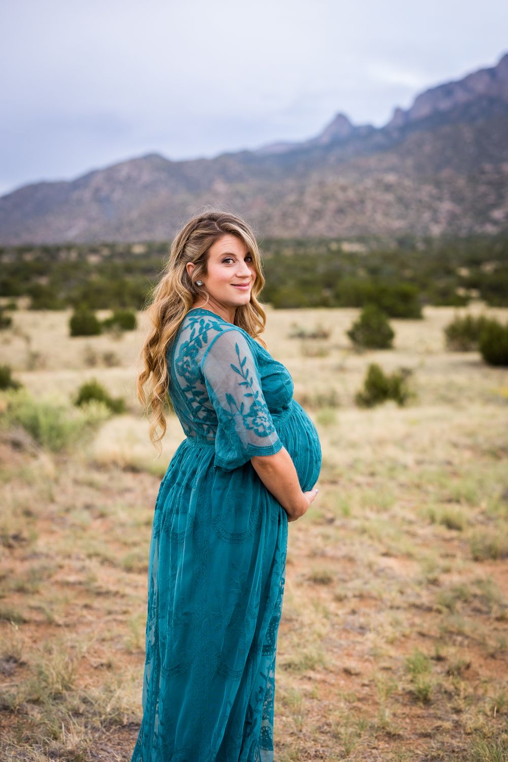 Expectant mother in teal lace dress poses against mountain landscape.