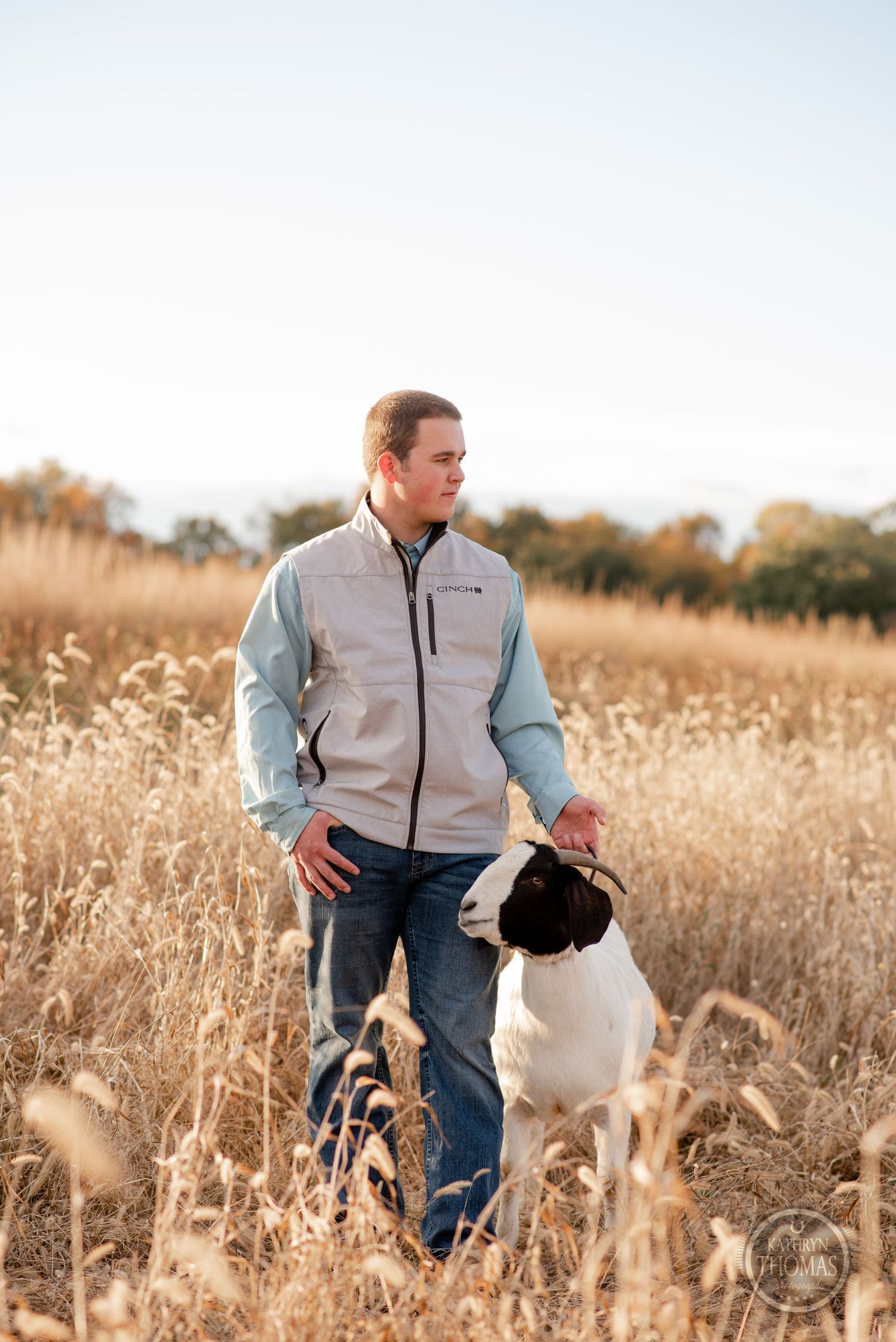 Person in vest and jeans stands in golden field with Boer goat at sunset.