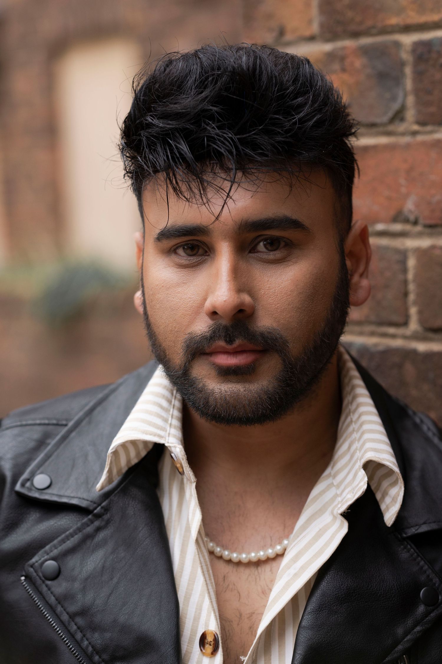 Stylish bearded man in a leather jacket and pearl necklace against a brick wall.