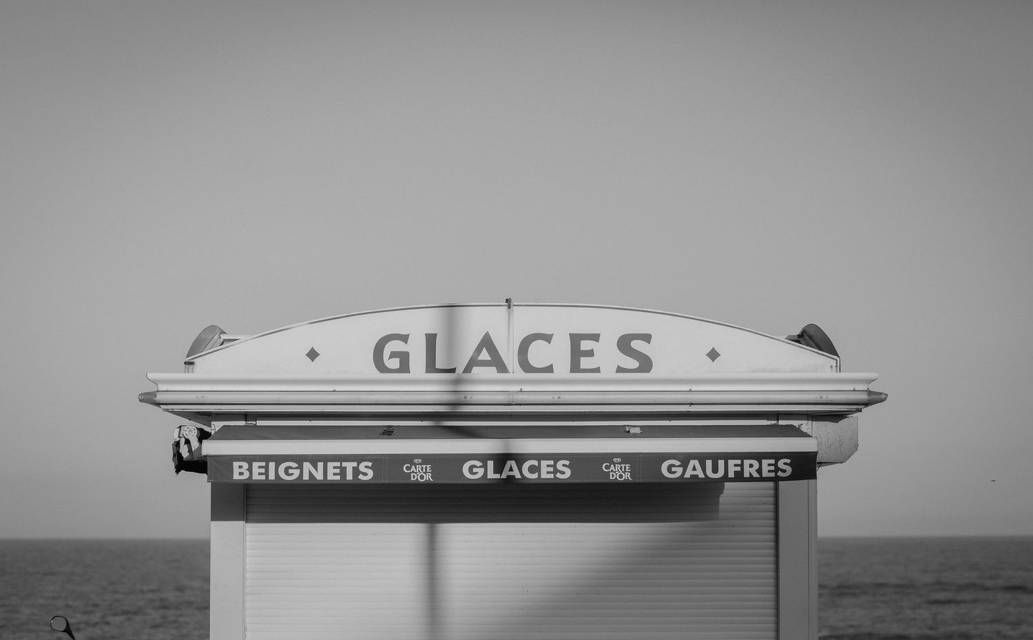 Black and white photo of a vintage ice cream stand sign reading 'GLACES' by the seaside.
