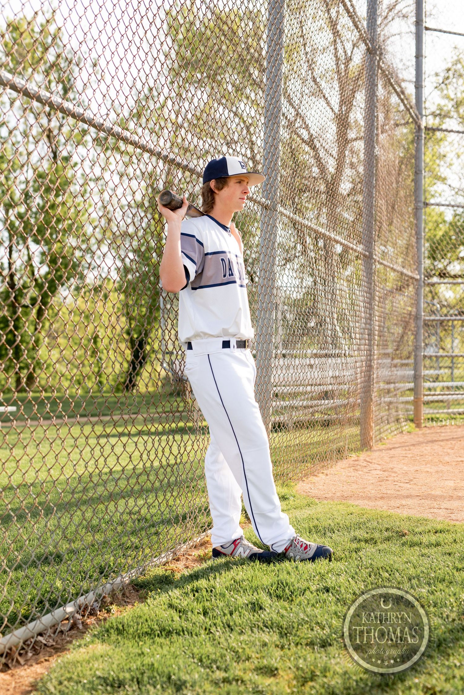 Baseball player in white uniform stands against fence during practice session.