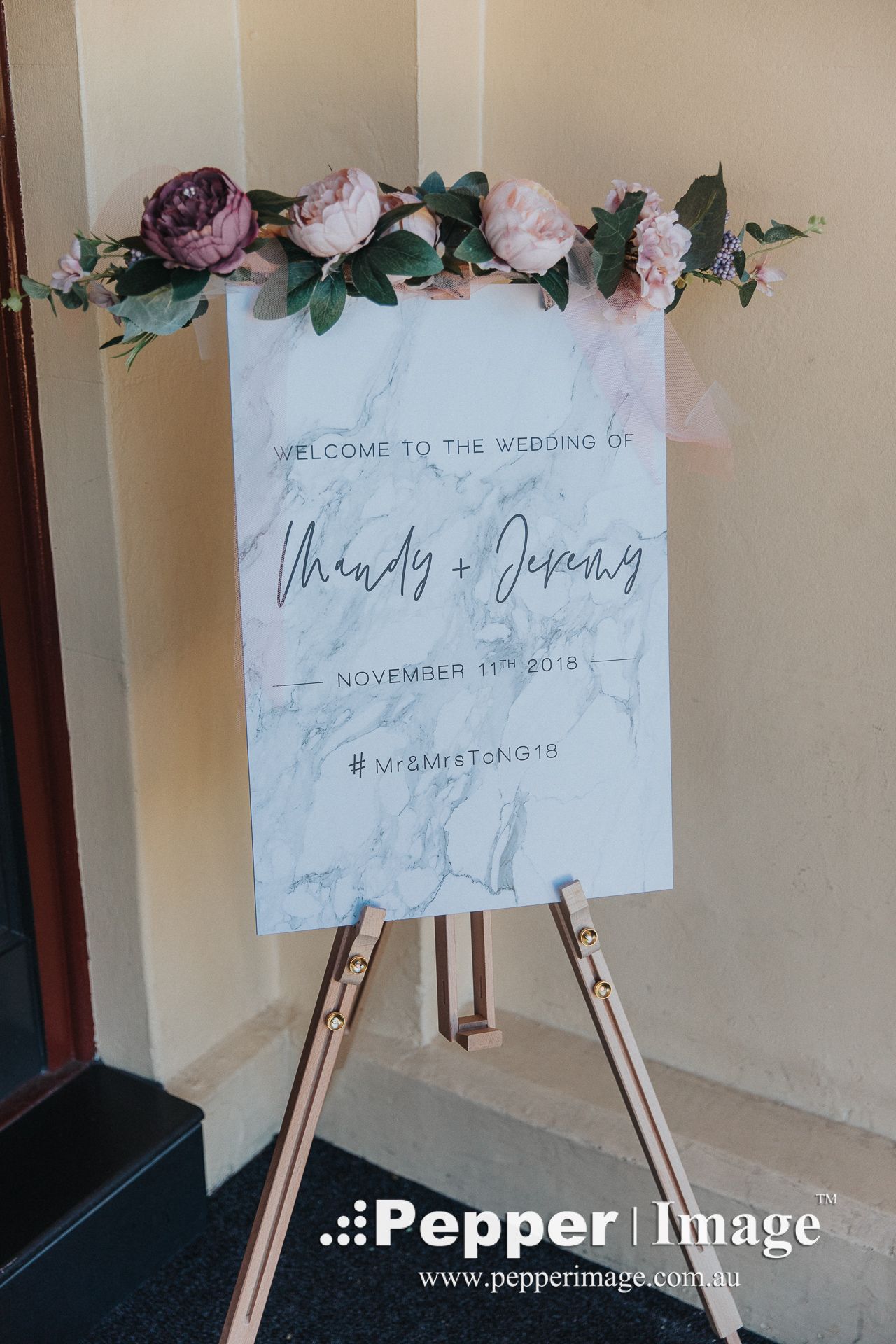 Elegant marble wedding welcome sign for Mandy and Jeremy decorated with pink and purple flowers on a gold easel.