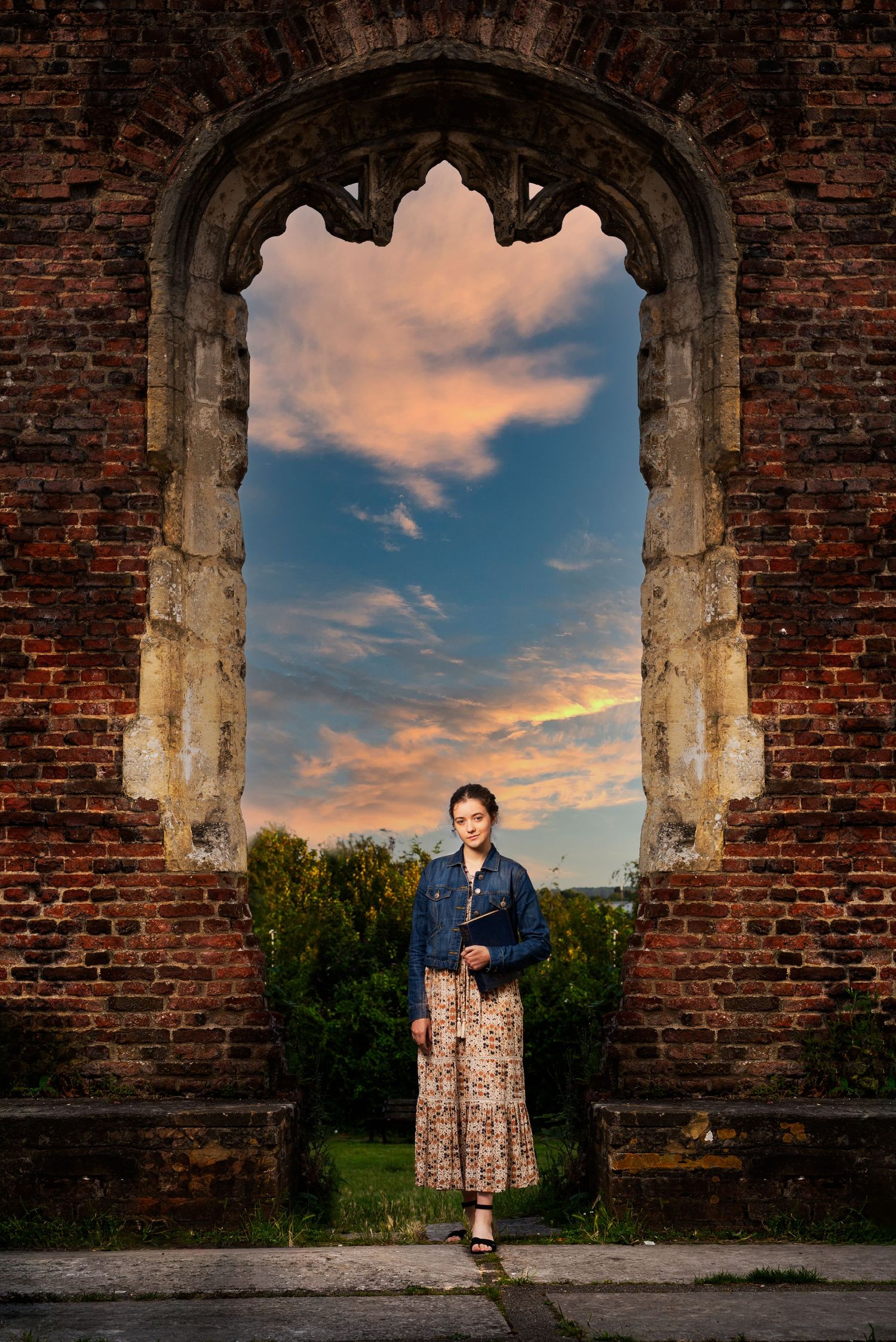 Atmospheric portrait through an old brick archway at sunset with dramatic orange clouds in the background.