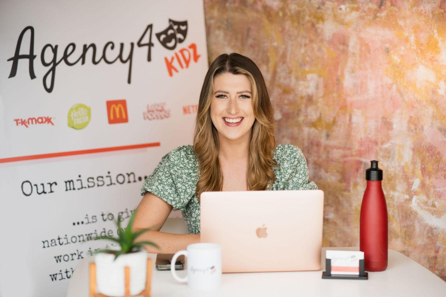 Woman in green floral top working on laptop at desk with Agency48 Kidz branding visible.