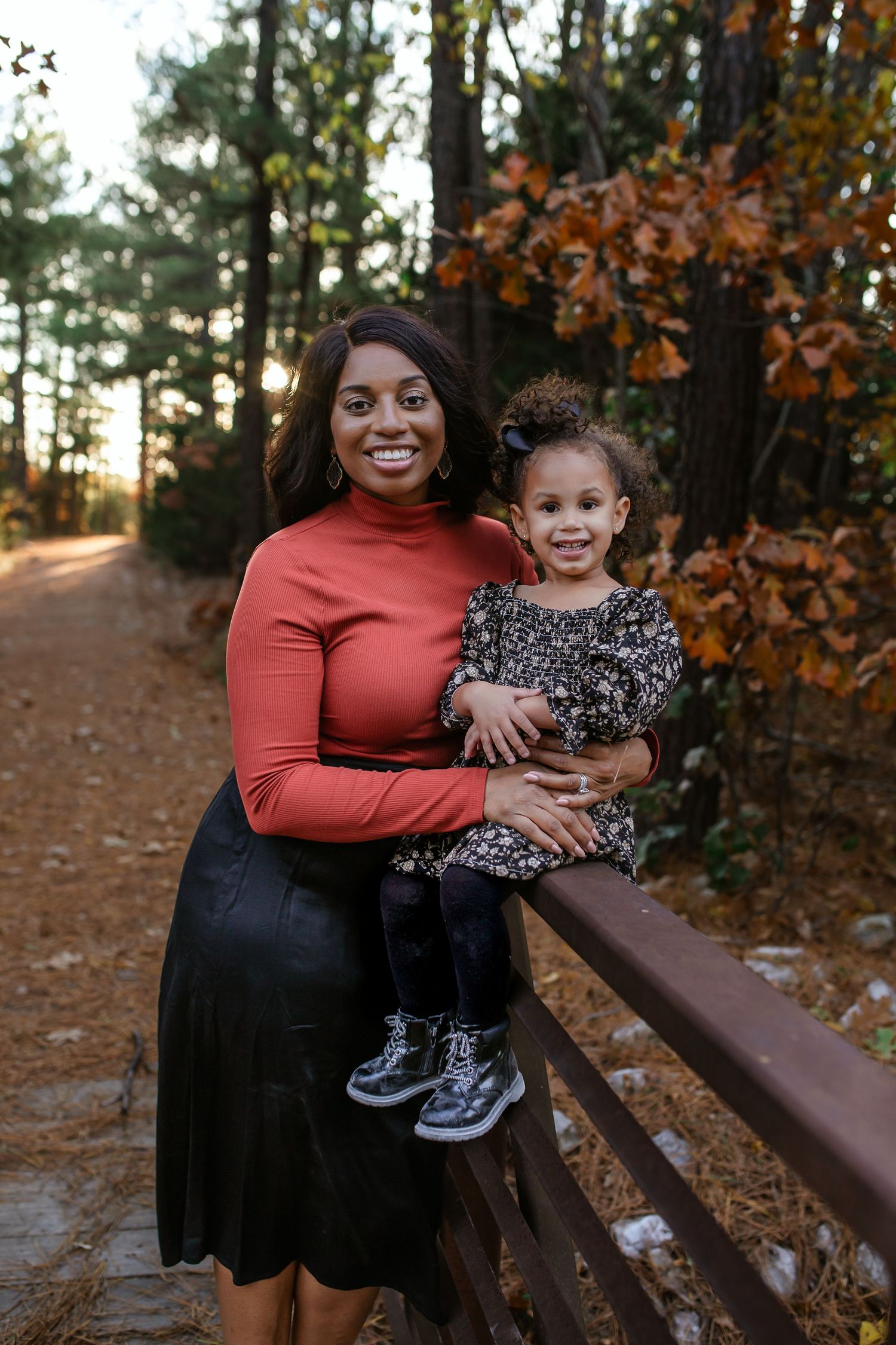 Mom and daughter pose on a bridge for fall family photos with bronze leaves to their right