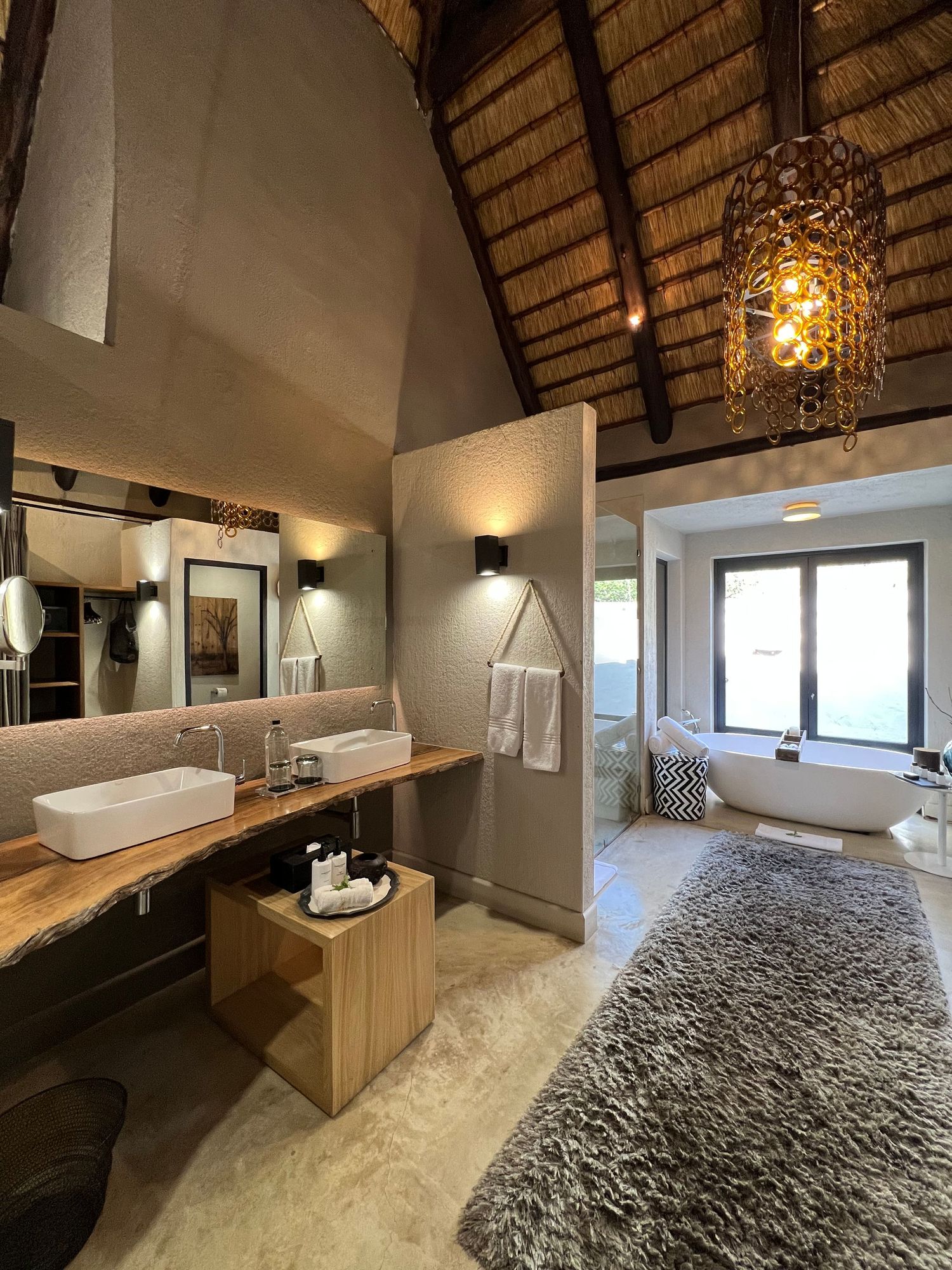 Luxurious safari lodge bathroom with thatched roof, twin vessel sinks, and modern amenities overlooking a spacious bathtub area.