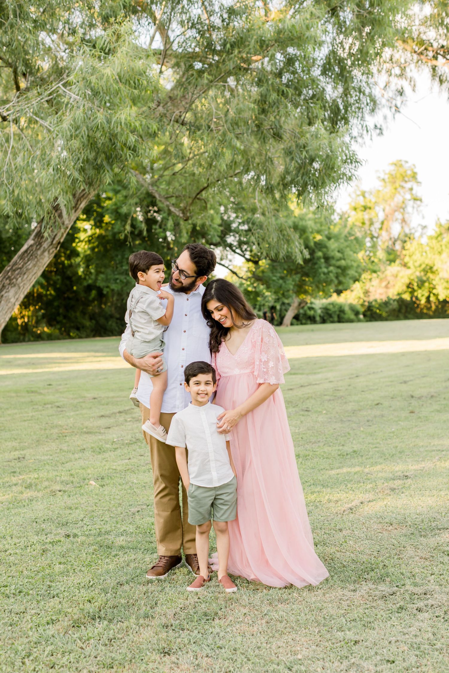 A family of four poses together under a willow tree in a park setting during golden hour.