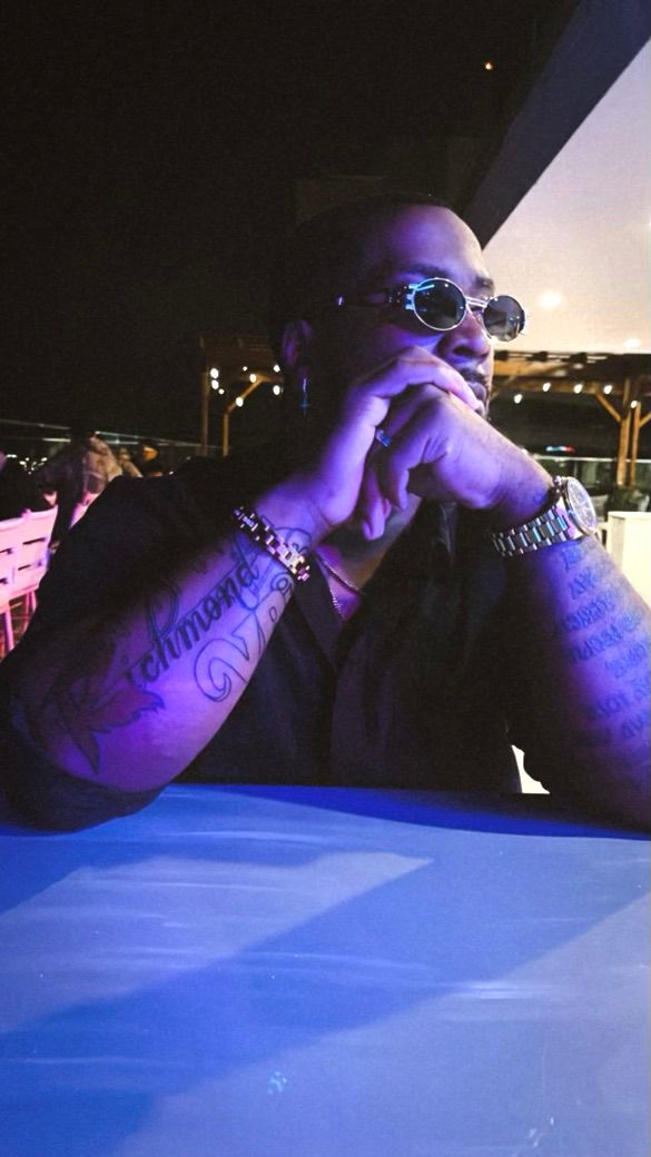 Person in sunglasses sits at table illuminated by blue lighting while wearing jewelry and showing tattoos.