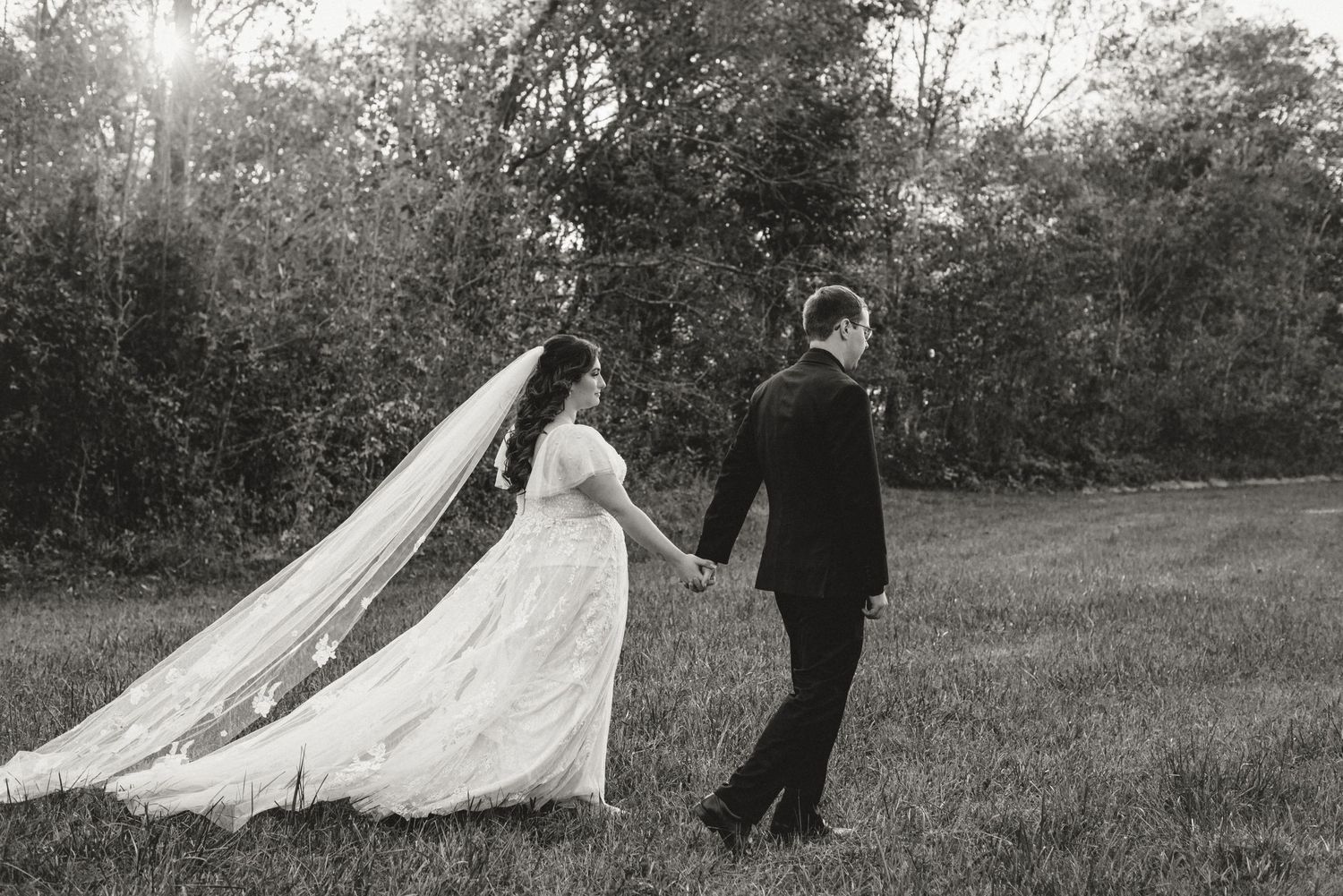 Romantic black and white photo of newlyweds walking hand in hand with flowing veil.
