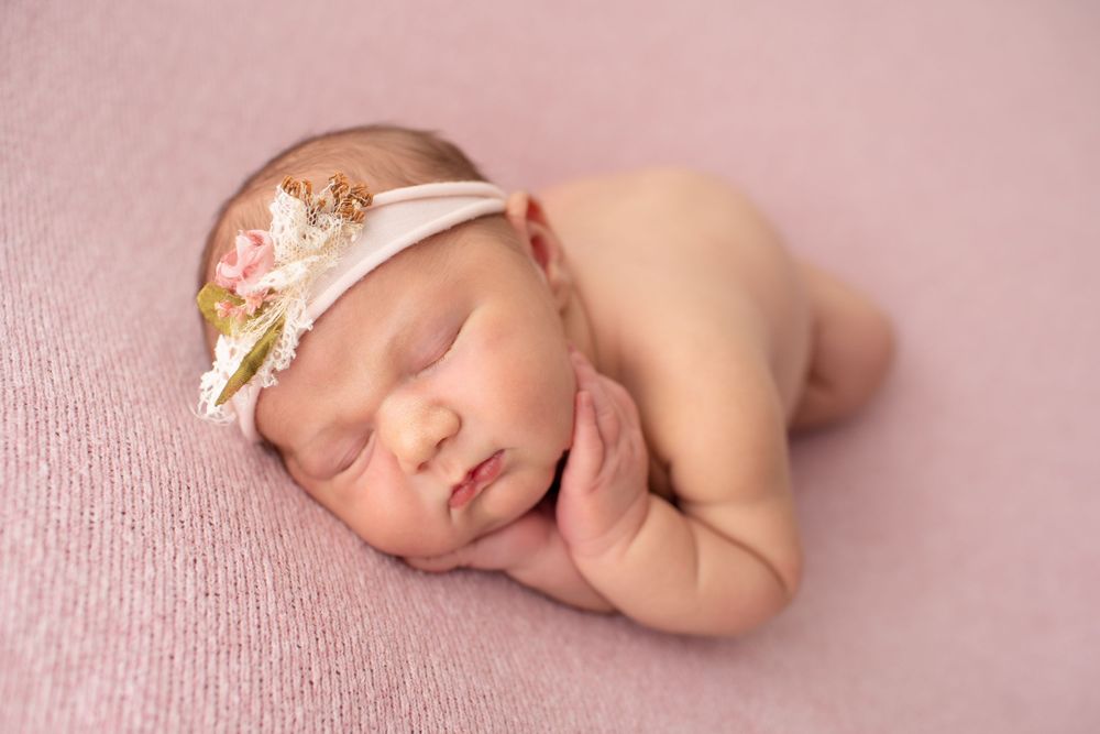 Peaceful sleeping newborn wearing floral headband during professional photo session on pink backdrop.
