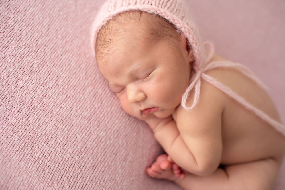 Newborn wearing soft pink bonnet sleeping on pink textured backdrop.