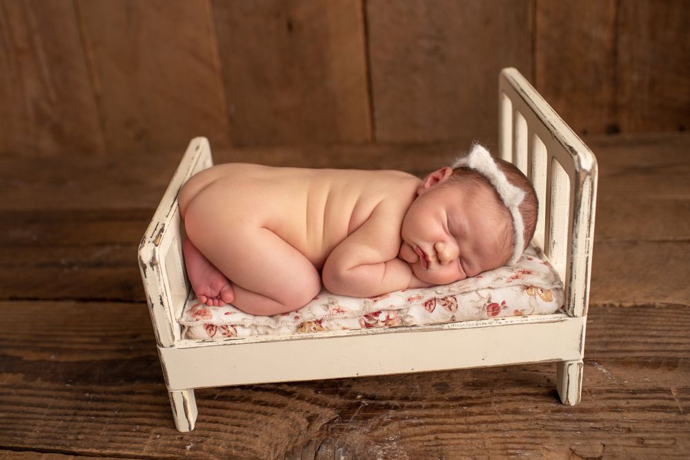 Sleeping newborn posed on vintage white wooden doll bed with floral bedding.
