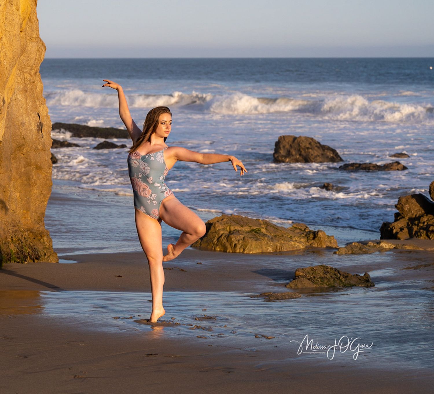 Dancer in gray leotard performs graceful pose on beach with crashing waves and rocky coastline.