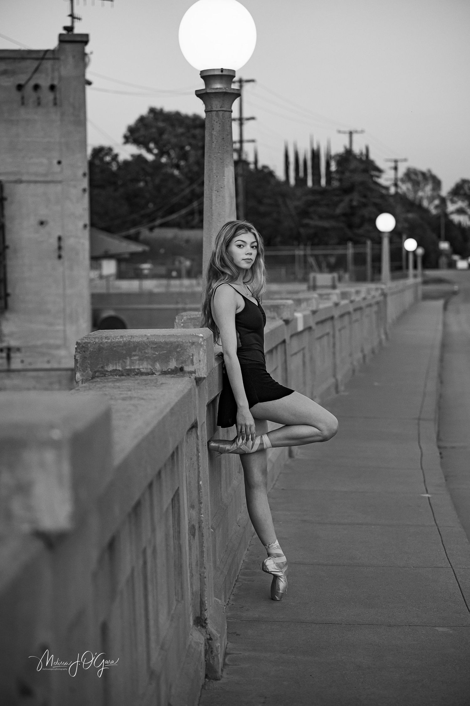 Black and white photo of ballet dancer posing en pointe along historic bridge railing.