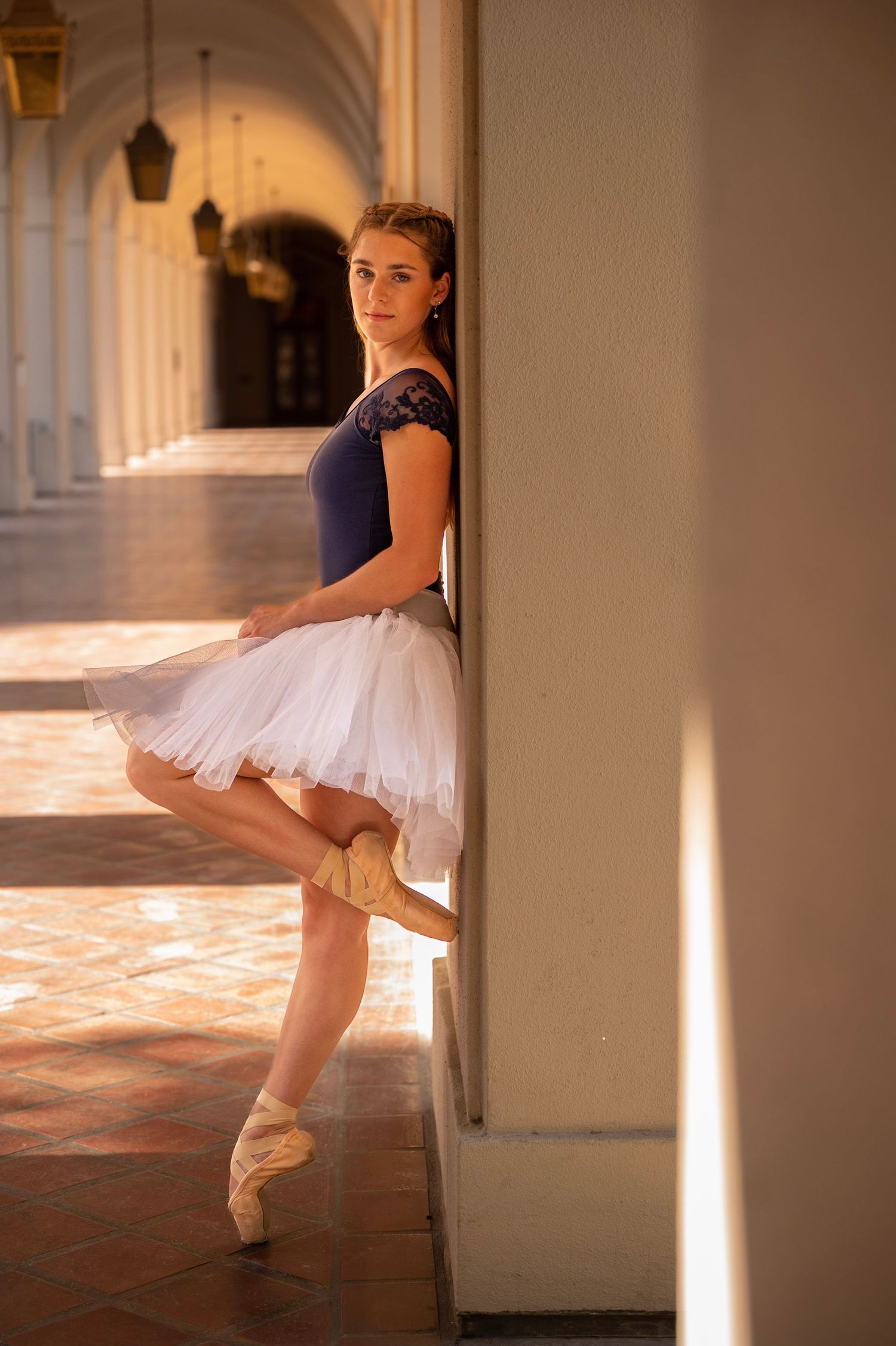 Ballerina in tutu and pointe shoes poses elegantly in arched corridor with dramatic lighting.