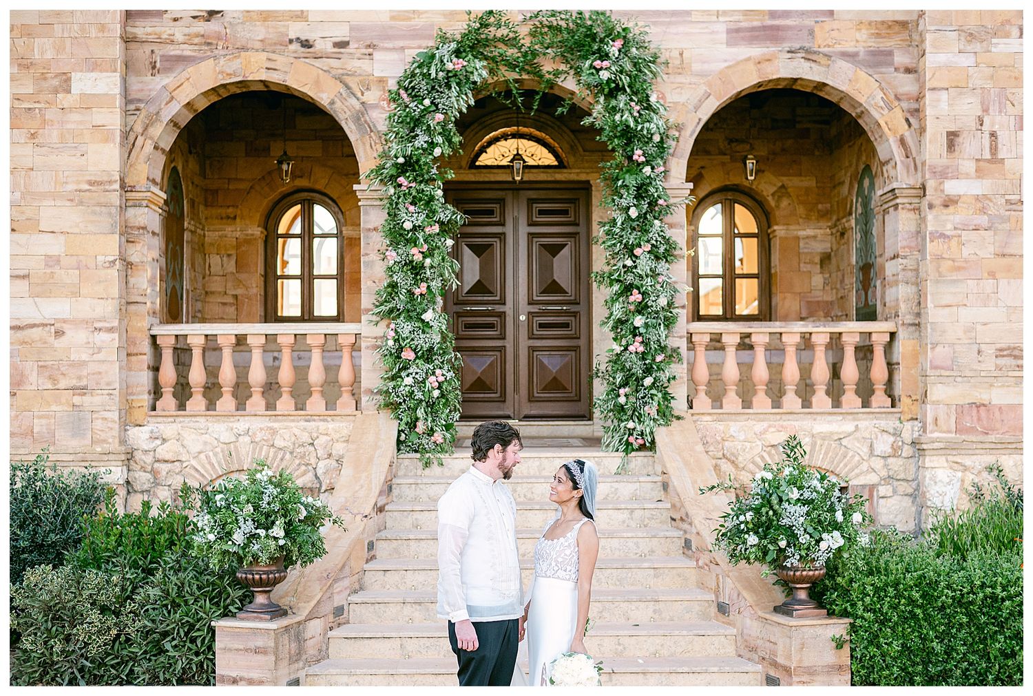 A romantic couple poses in front of a Mediterranean stone archway adorned with climbing vines and flowers.
