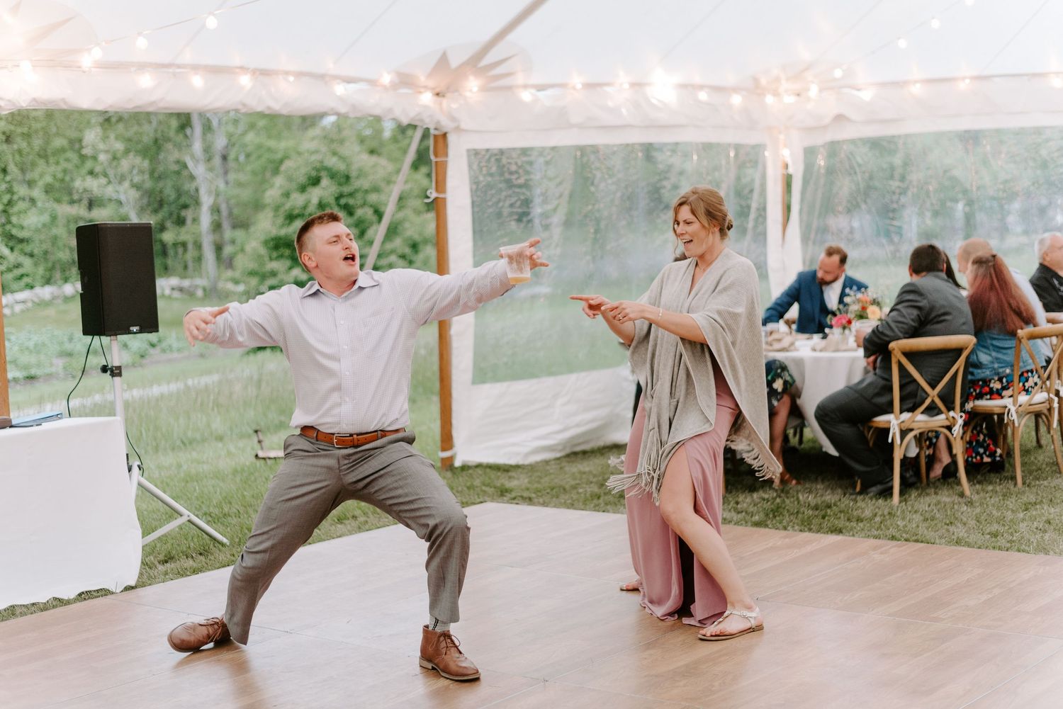 Wedding guests dancing enthusiastically on a wooden dance floor under a tent.