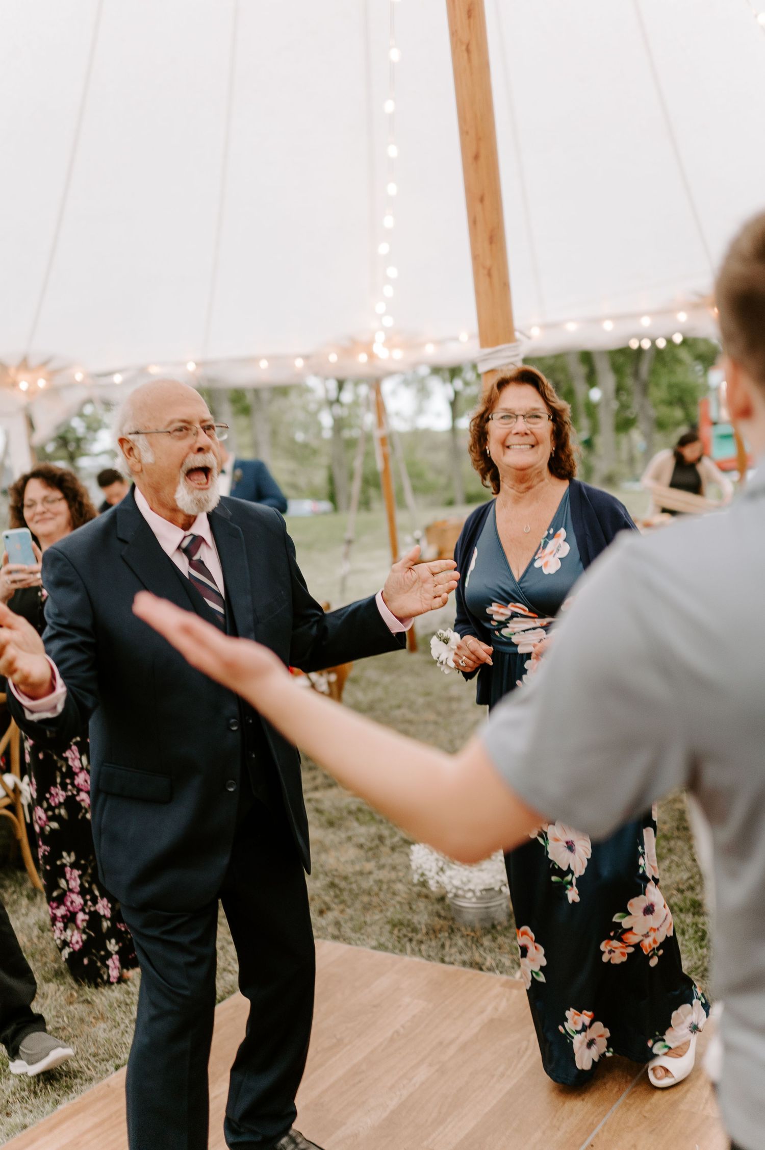 Wedding guests dancing in formal attire under a tent with string lights at dusk.