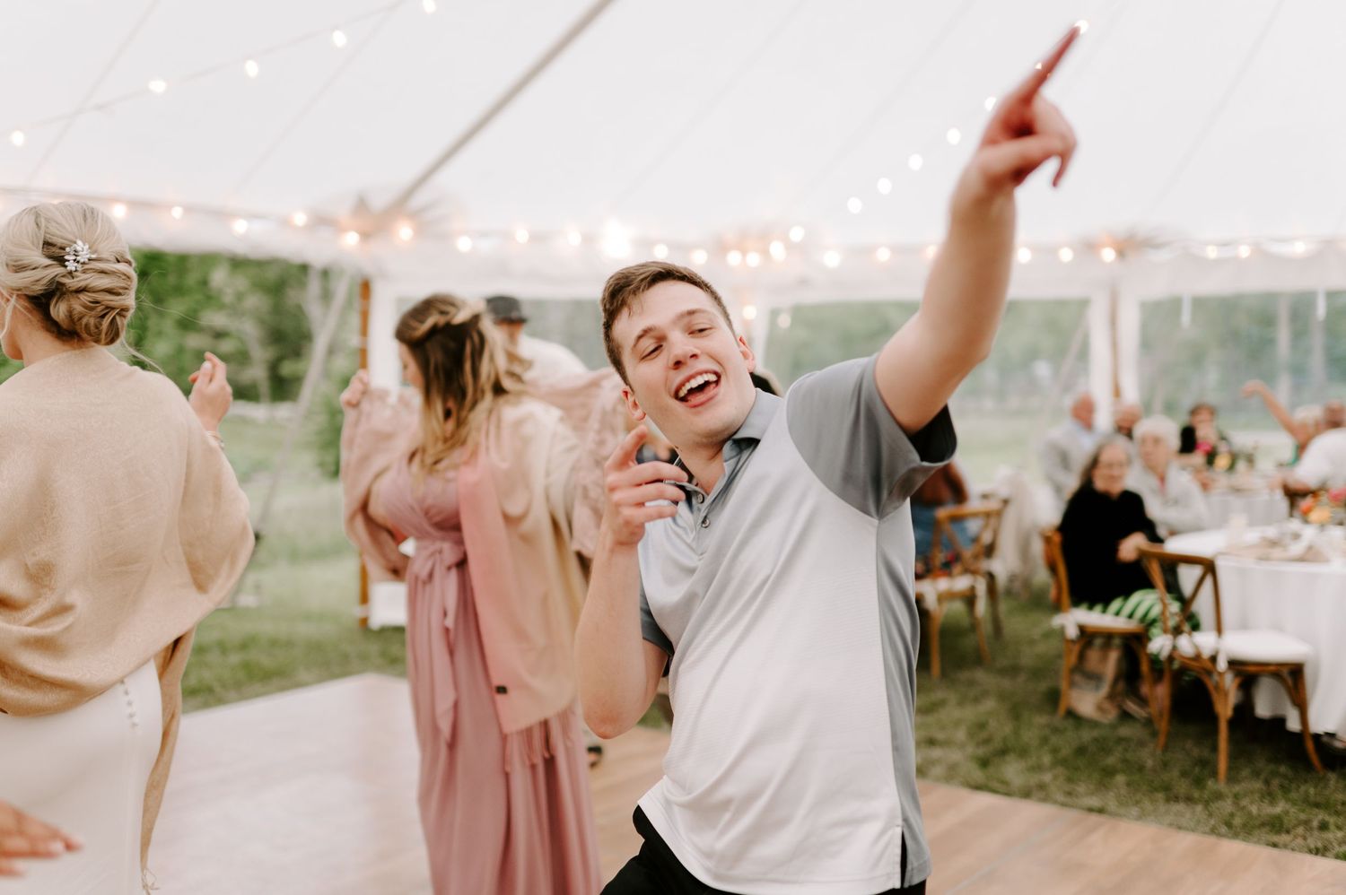 Wedding guests dancing and celebrating under a white tent with string lights at an outdoor reception.