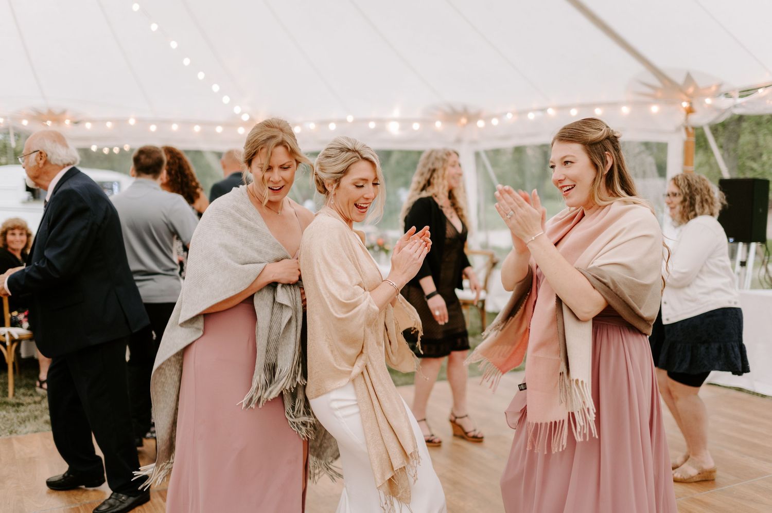 Wedding celebration with guests dancing and laughing under a tent decorated with string lights.