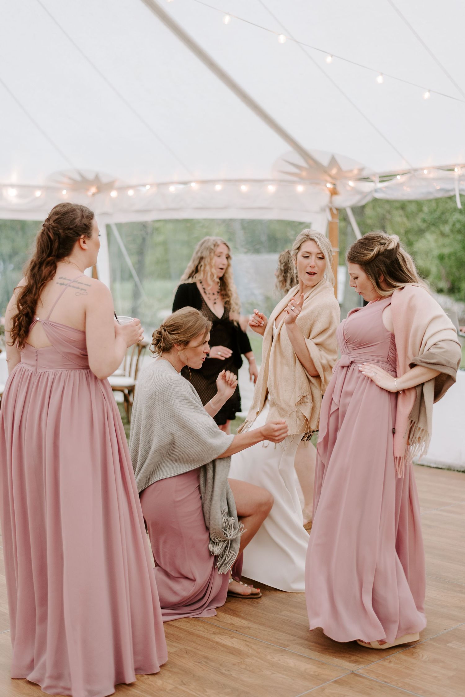 Wedding party in pink bridesmaid dresses dancing and celebrating under a tent with string lights.