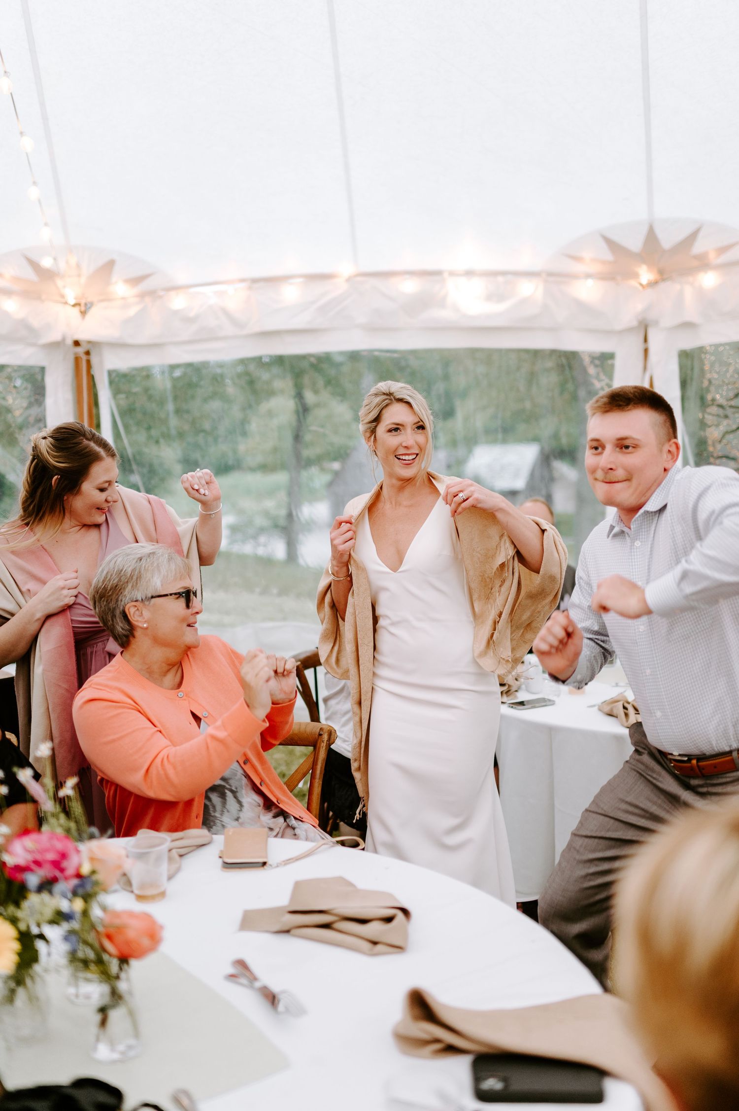Wedding reception dinner setup with string lights and floral centerpieces under a white tent.