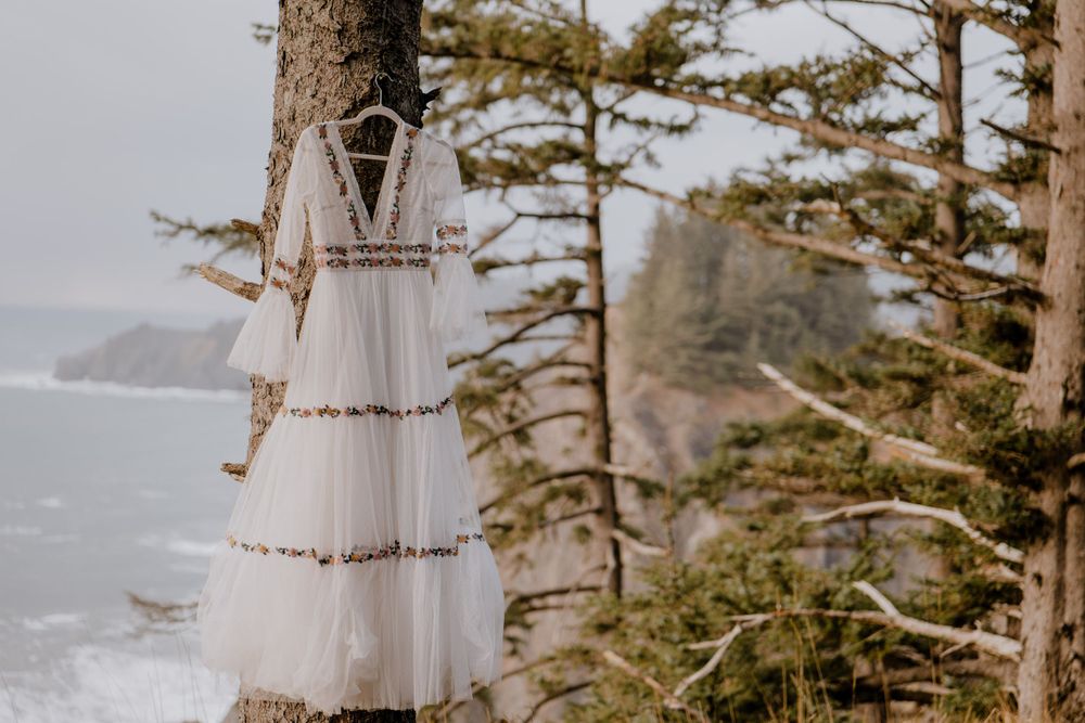 White wedding dress with floral accents hung on pine tree overlooking ocean