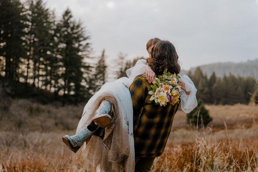 Groom carrying bride in grassy field surrounded by trees
