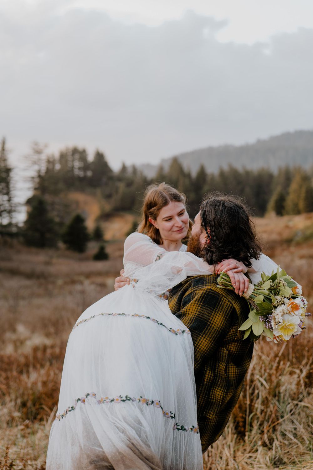 Groom carrying bride in grassy field while bride looks at him