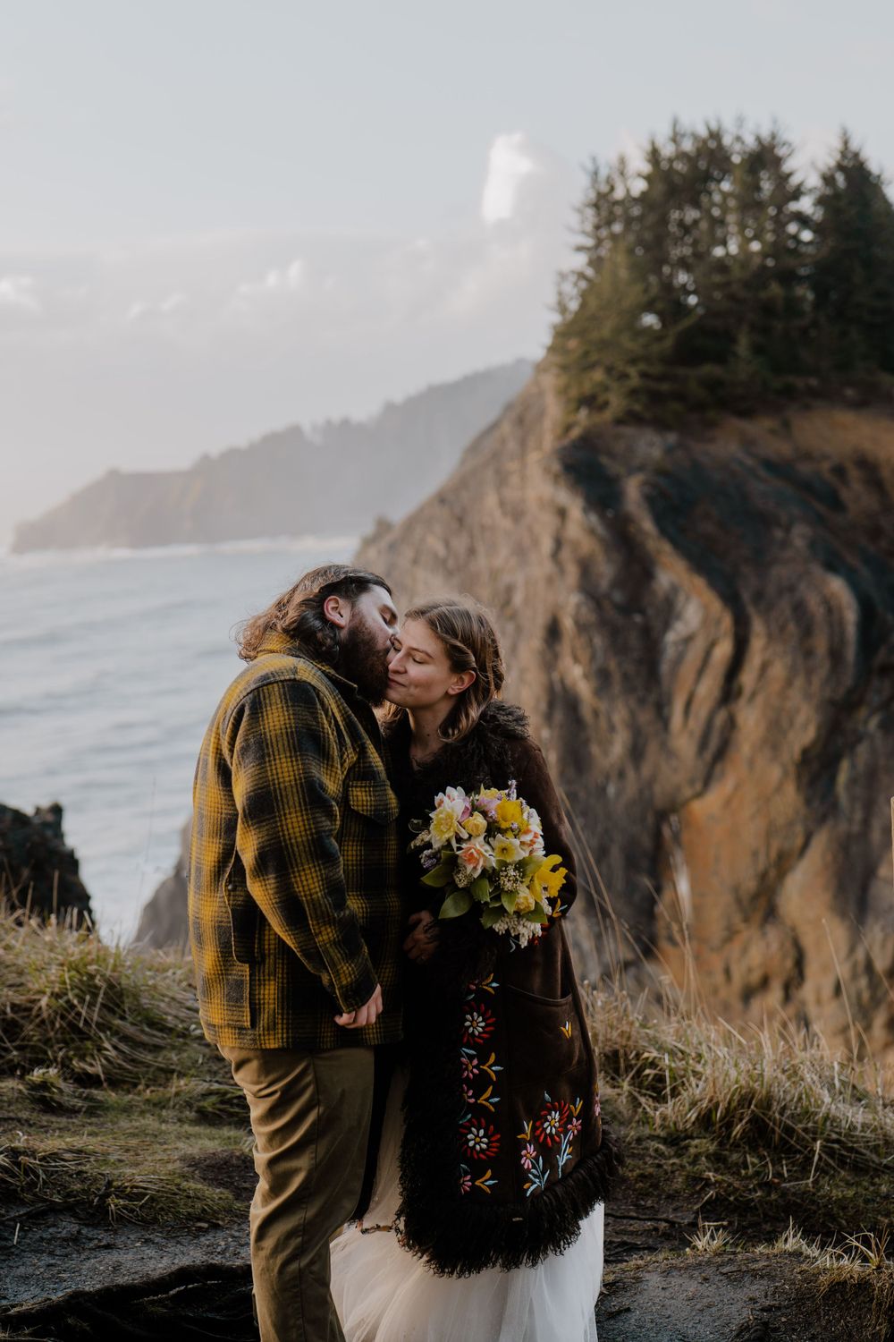 Groom kissing bride on the cheek next to coastal cliffside