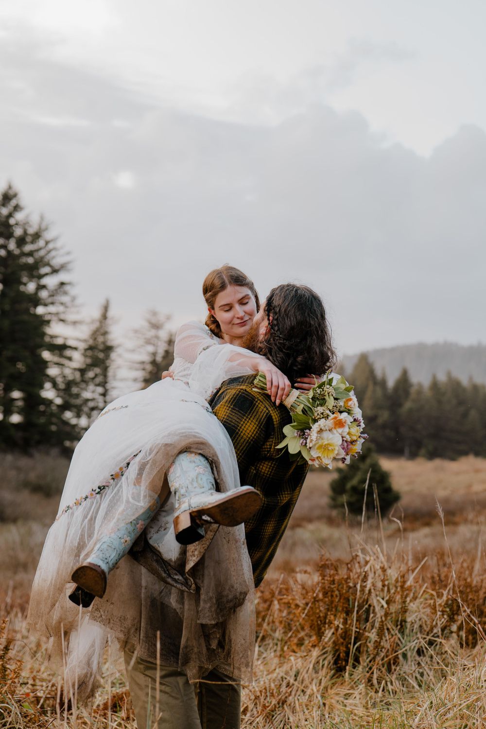 Groom carrying bride in grassy field while bride looks at him