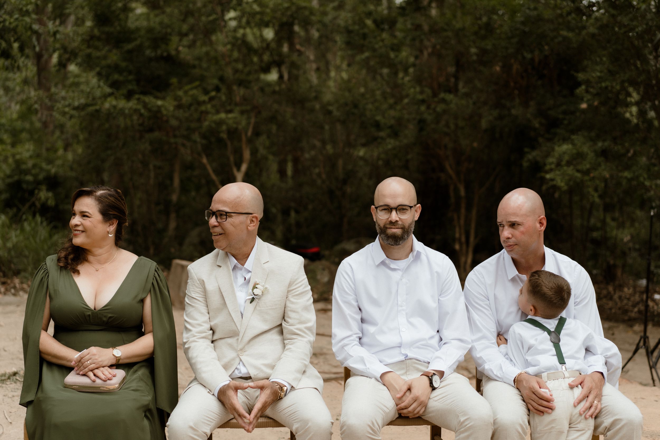 Wedding guests in coordinated outfits seated on wooden benches in a forest setting.
