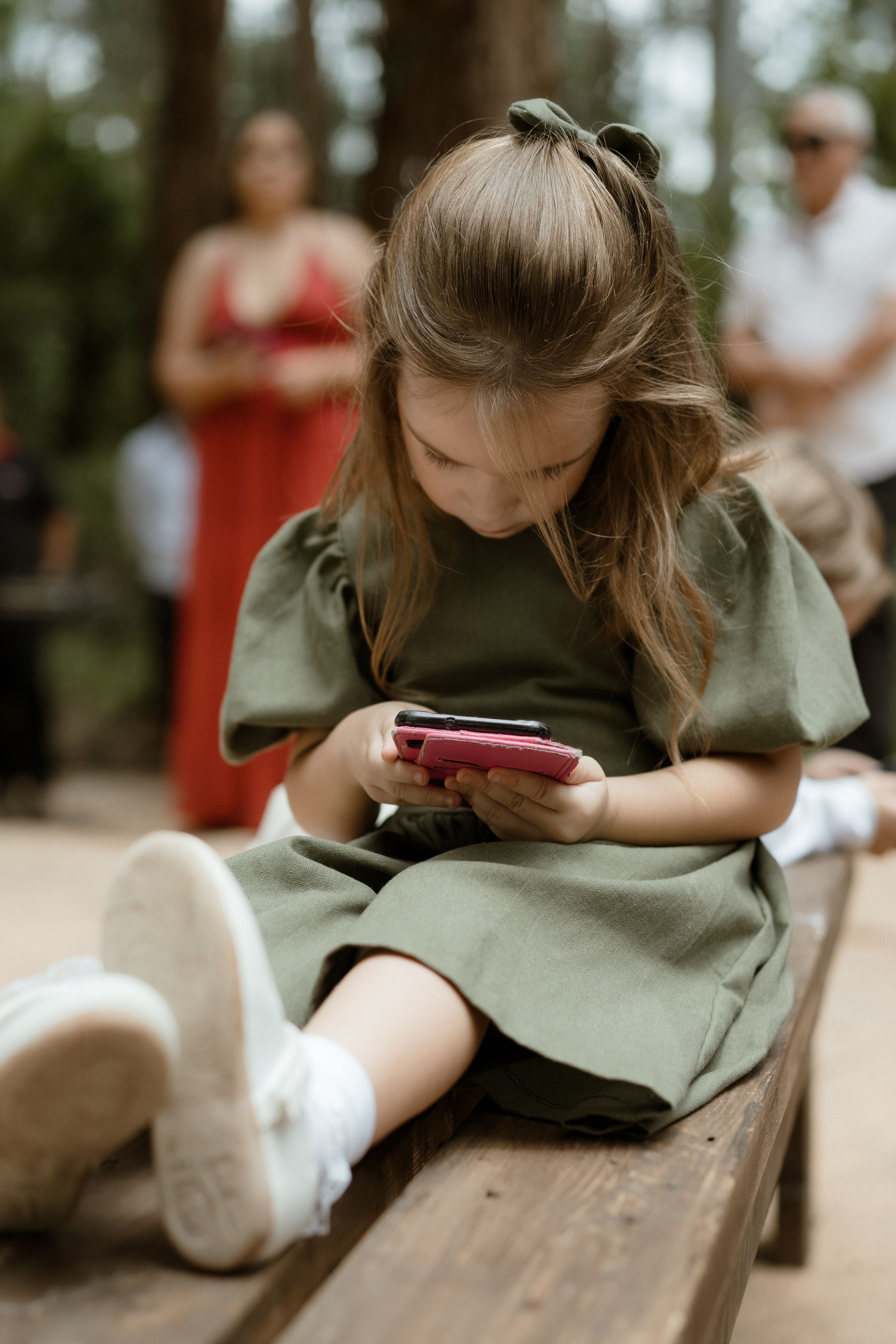 Young attendee in olive green dress uses phone while sitting on wooden bench outdoors.