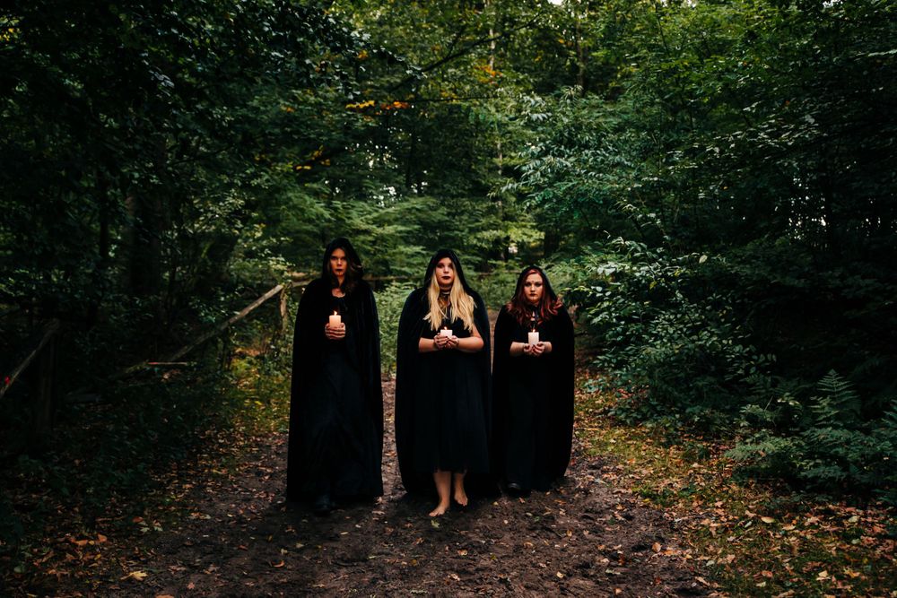 Three people in black robes hold candles while standing on a wooded path.